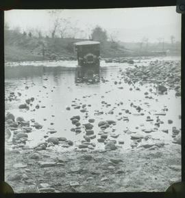 Thornycroft bus fording river with stony bed.