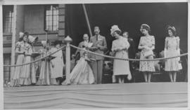 Young woman wearing a Voortrekker bonnet presenting something to the Royal family on a dais.