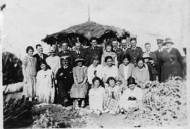 Uniformed men with civilian women posing before simple thatched rondavel.