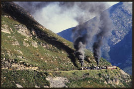 Two steam locomotives with passenger train in mountain pass.