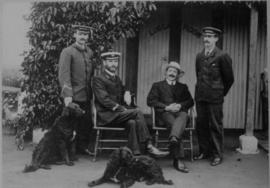 Four men and two dogs in front of corrugated iron luggage room.