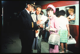 Conductor checking passenger list with passengers on station platform.