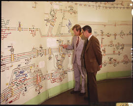 
Two men studying schematical railway layout on wall in control room.
