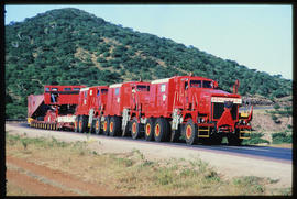 
Heavy load hauled by three motor trucks on road.
