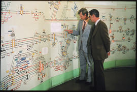 
Two men studying schematical railway layout on wall in control room.
