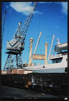 Harbour crane transferring large wooden crates between train and ship.