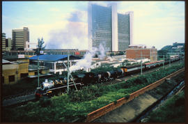 
SAR Class 19D steam locomotive with torpedo tender on passenger train in city.

